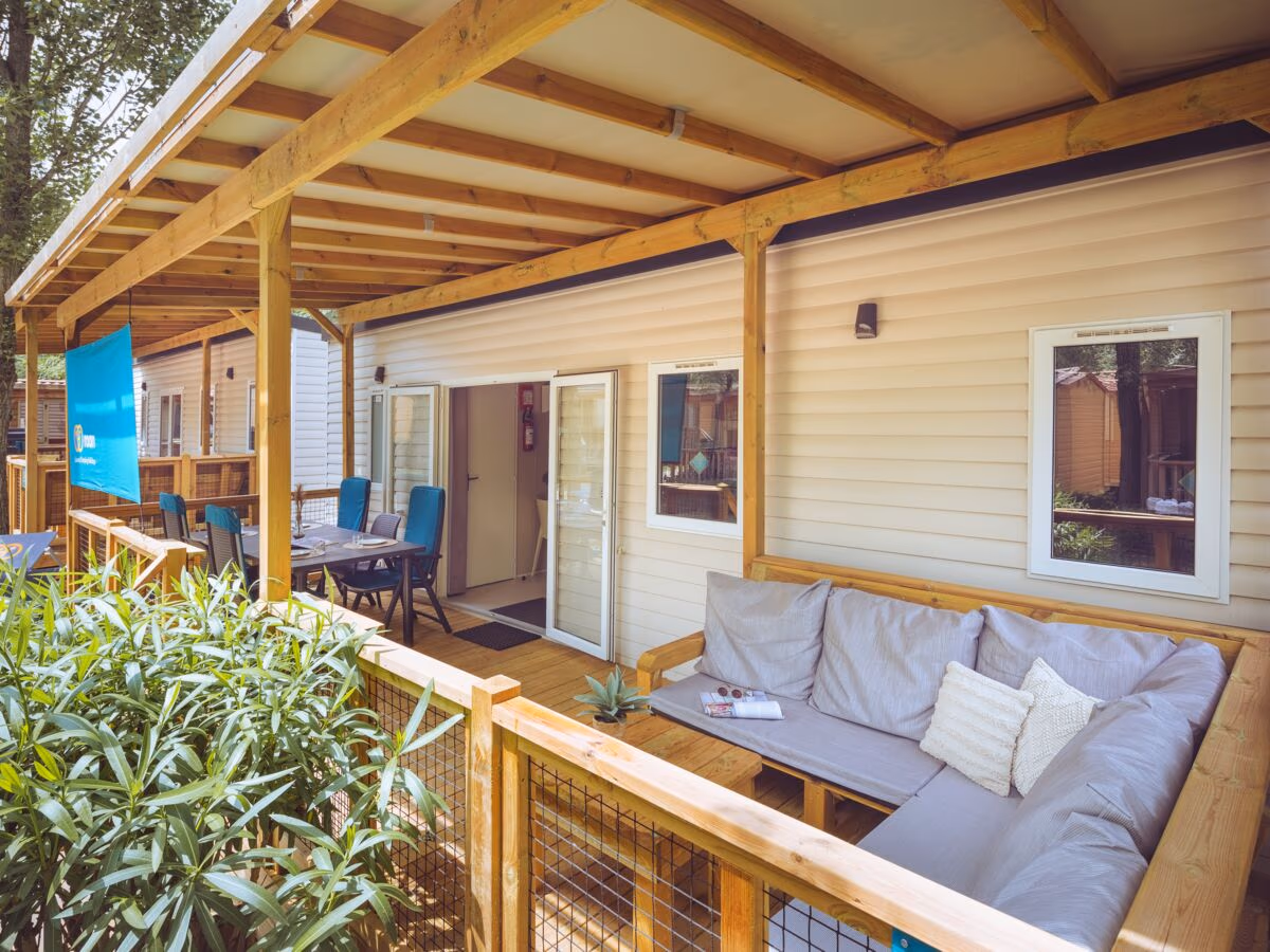Veranda onder een houten balkenkap met een hoekbank en verderop een eettafel, bij een stacaravan