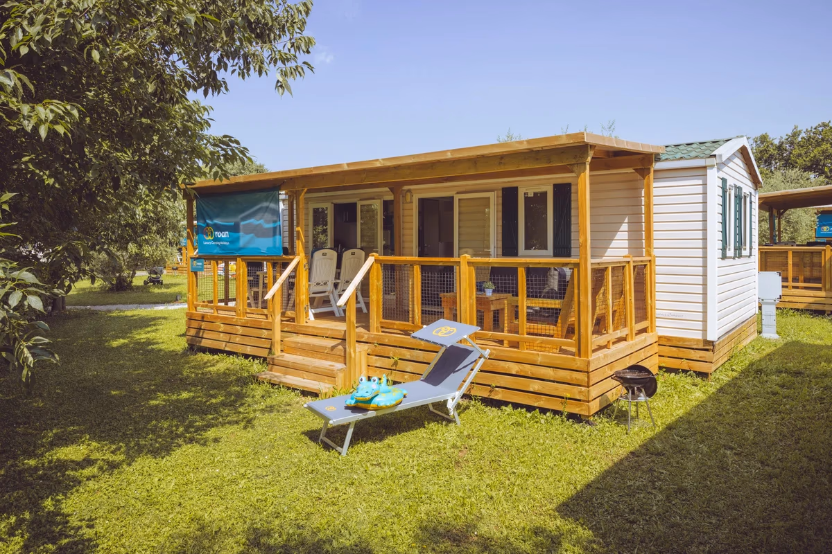 Stacaravan met een houten overkapping en veranda in het gras op Camping Bijela Uvala, met een ligbed en slippers ervoor