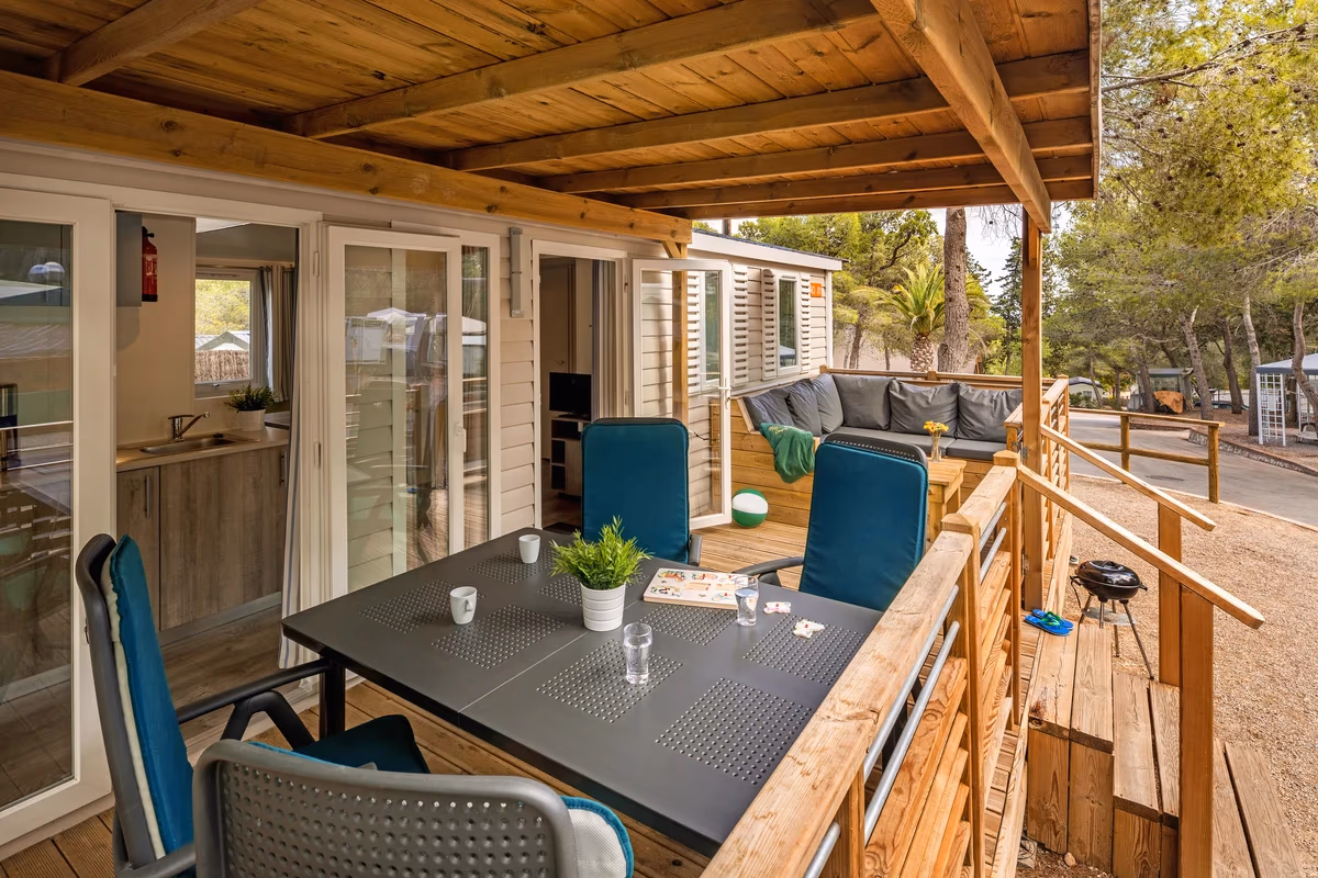 Overdekt terras met een eettafel en blauwe stoelen bij een stacaravan op Camping Bijela Uvala, met doorkijk naar de keuken