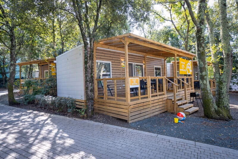 Stacaravan met houten gevel en een overdekt terras onder hoge bomen op Camping Bijela Uvala, met een gele vlag aan de overkapping