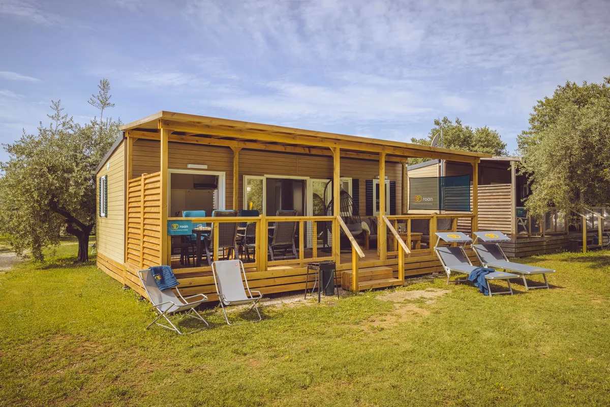 Houten stacaravan met een overdekte veranda op Camping Bijela Uvala, met ligbedden en klapstoelen in het gras ervoor