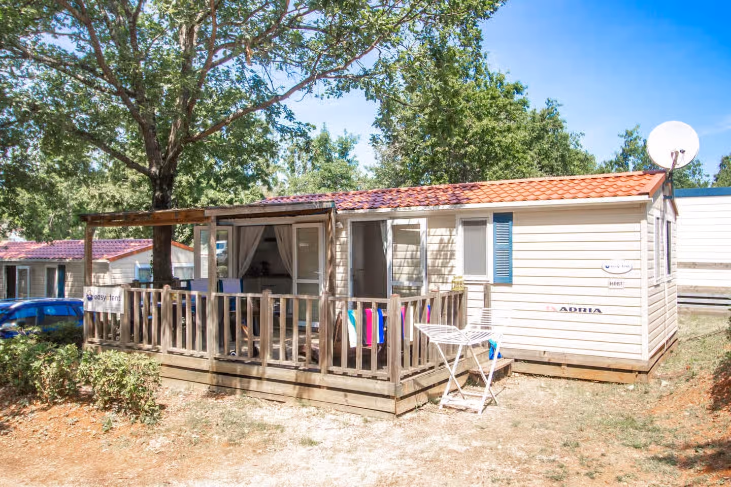 Stacaravan met blauwe luiken, een rood dak en een houten terras op Camping Bijela Uvala, met een satellietschotel aan de zijkant