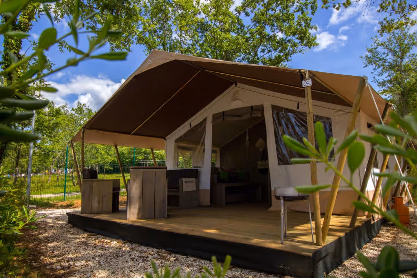 Safaritent met een donkere luifel op houten palen en een houten terras met loungebanken, tussen het groen onder een blauwe lucht