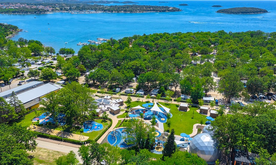 Luchtfoto van het beboste terrein van Camping Bijela Uvala met speelplekken en een witte koepeltent, en daarachter de baai met eilandjes