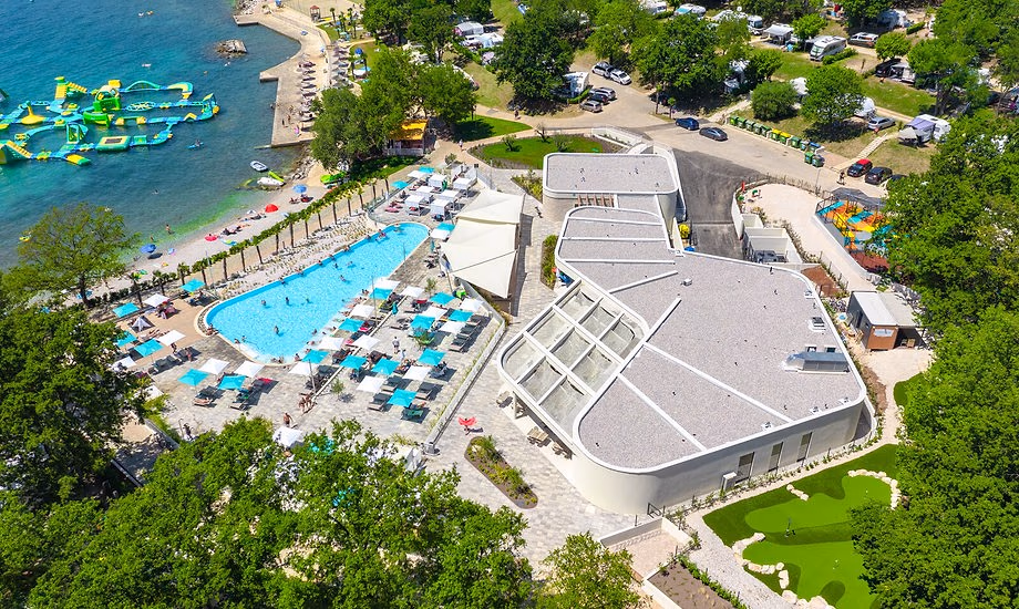 Luchtfoto van het zwembad met turquoise parasols aan de kust van Camping Bijela Uvala, met het opblaasbare waterpark in zee en een gebouw met een golvend wit dak