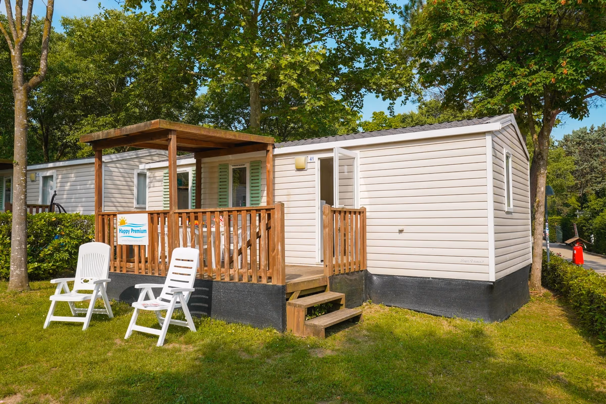 Witte stacaravan met een houten veranda en een naambordje Happy Premium, met twee witte stoelen in het gras