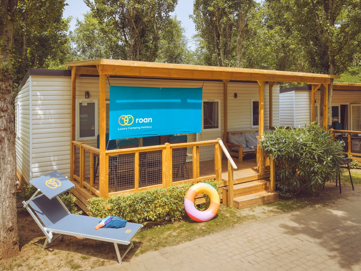 Stacaravan met een brede houten overkapping en een doek als zonwering, met een ligbed en een zwemband ervoor