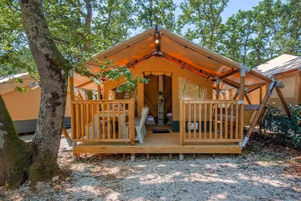 Lodgetent met houten veranda en openstaande voorzijde tussen de bomen op een bosrijke camping